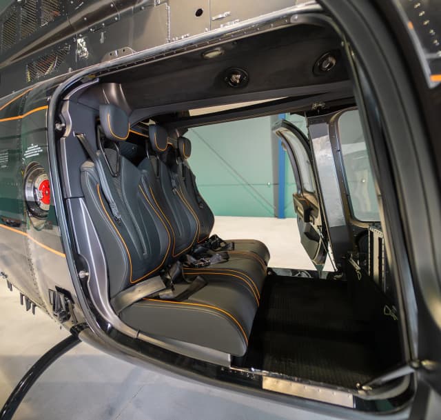 Row of dark Bell 407 passenger seats with orange piping and shoulder belts, viewed through the cabin doorway.
