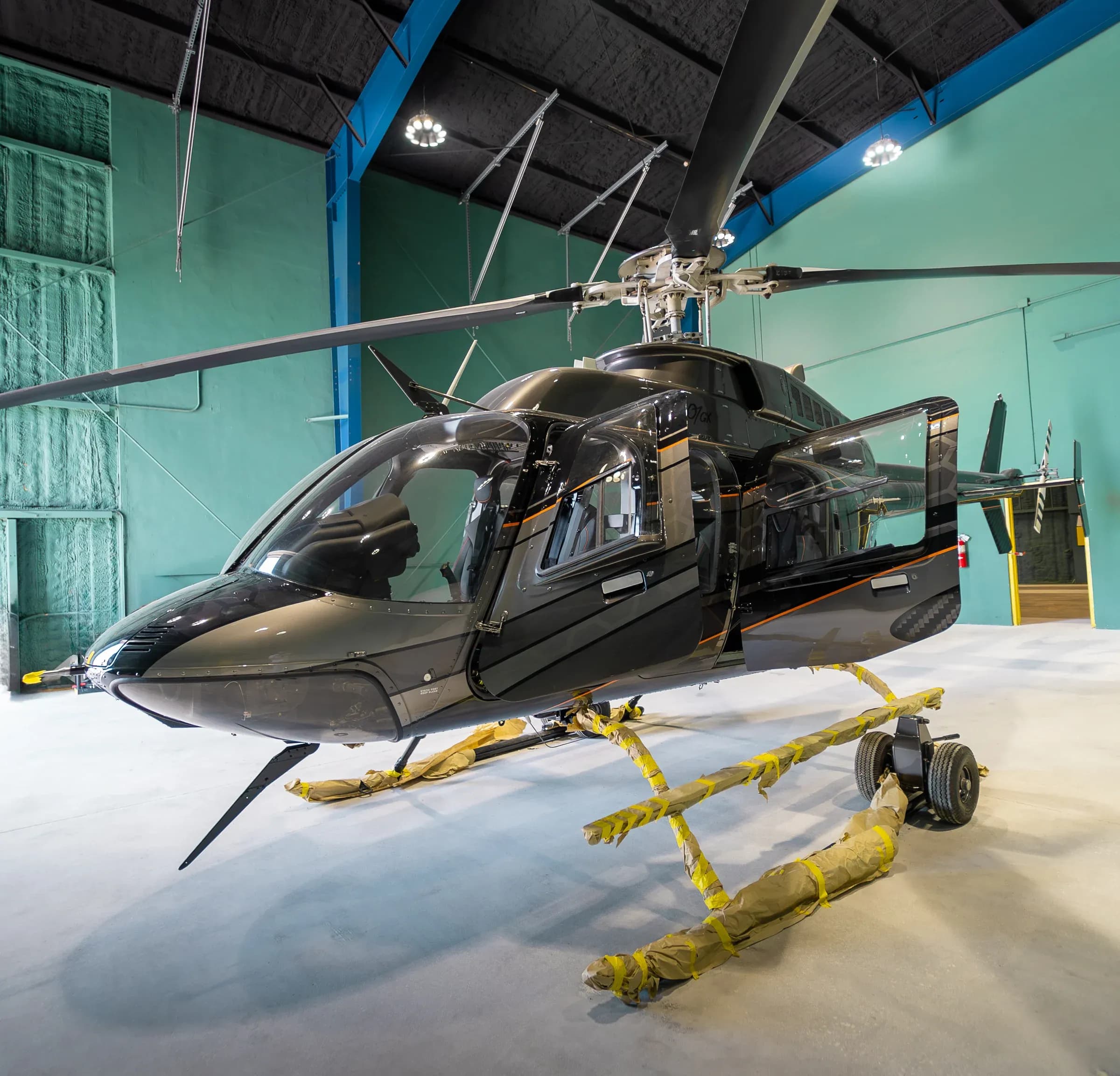Front-side view of the dark Bell 407 inside a hangar with doors open and ground handling equipment.