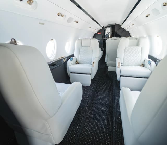 Centered view through the Gulfstream G200 cabin, with white diamond-quilted seats and dark carpet.