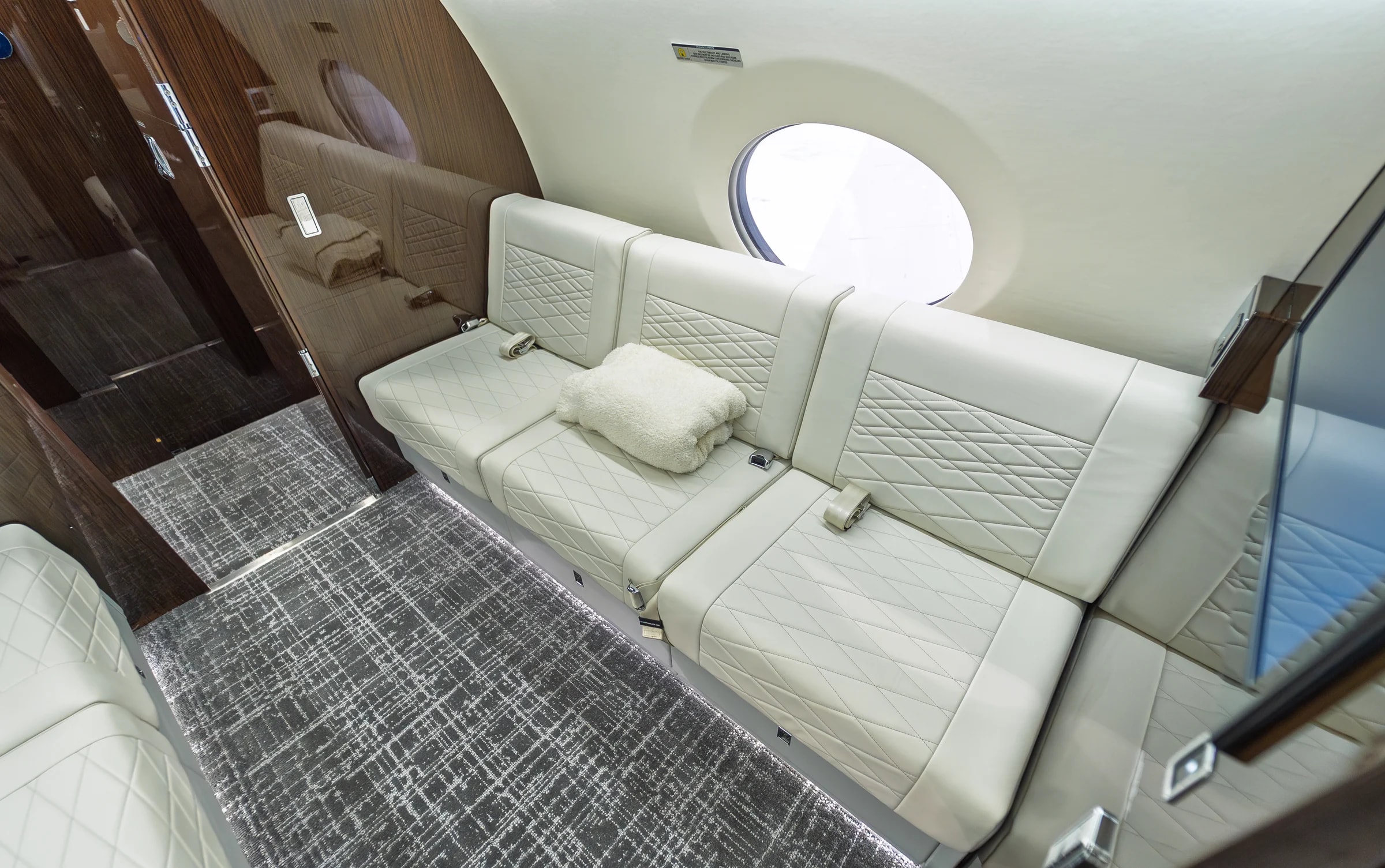 Gulfstream G650ER ivory divan with geometric quilting, a folded cream blanket, glossy wood-toned trim and patterned carpet.